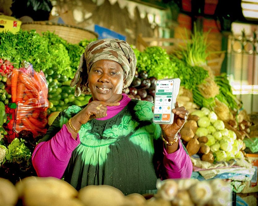 Commerçante souriante devant son étal de légumes, présentant un terminal SamirPay